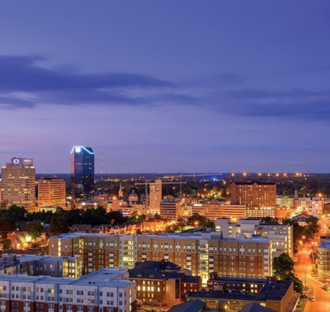 A panoramic view of the downtown Lexington, Kentucky skyline at dusk, featuring tall buildings illuminated against a colorful evening sky, with greenery and urban structures in the foreground.