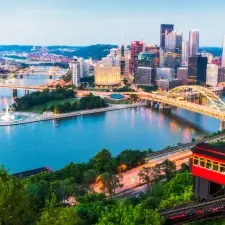 A scenic view of downtown Pittsburgh, showcasing the city skyline, rivers, and bridges under a bright blue sky. The image captures the beauty and architectural landmarks of the city, including prominent high-rise buildings and lush green surroundings.