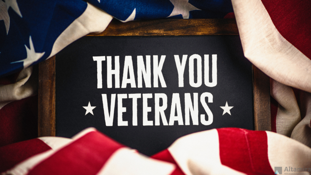 A chalkboard sign with the message ‘THANK YOU VETERANS’ written in bold white letters, framed by a backdrop of American flags in red, white, and blue, conveying appreciation for military service.